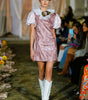 Model walking on a fashion show runway wearing a pink dress and white boots.