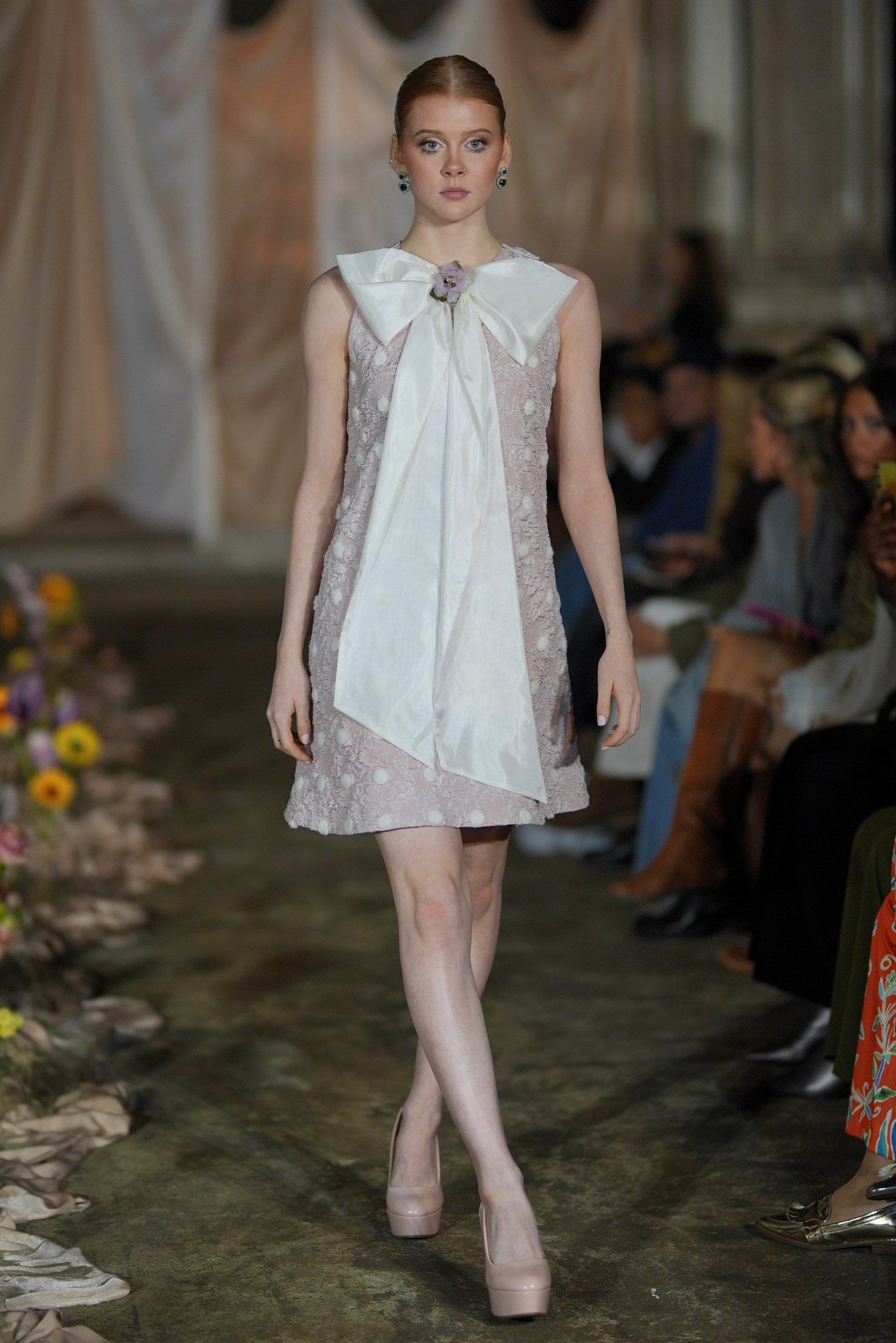 Model walking on a runway wearing a floral dress with a large bow at the front.