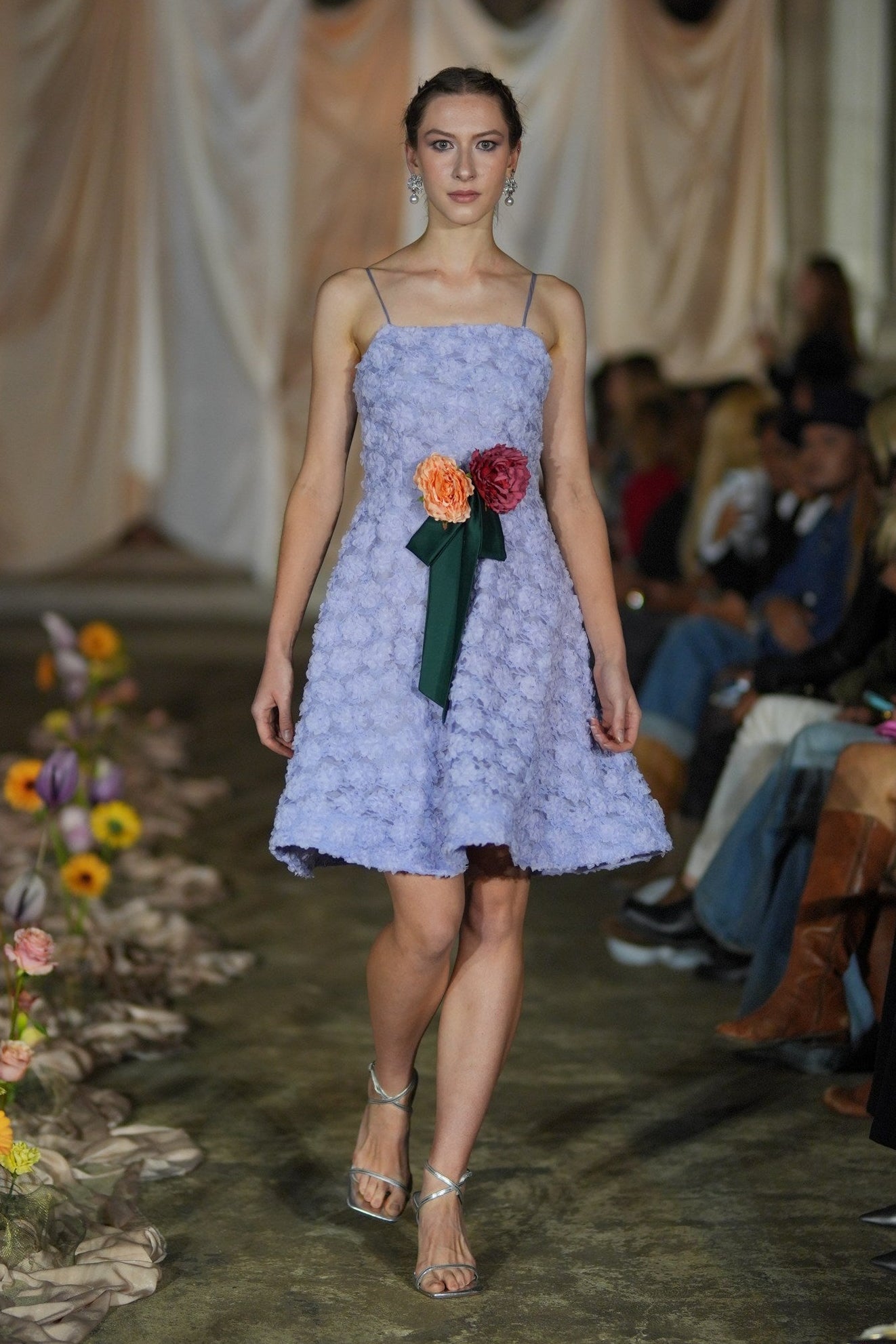 Model wearing a light purple dress with floral accessories on a runway.