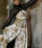 Model wearing a black sequined top and white floral pants against a textured wall.