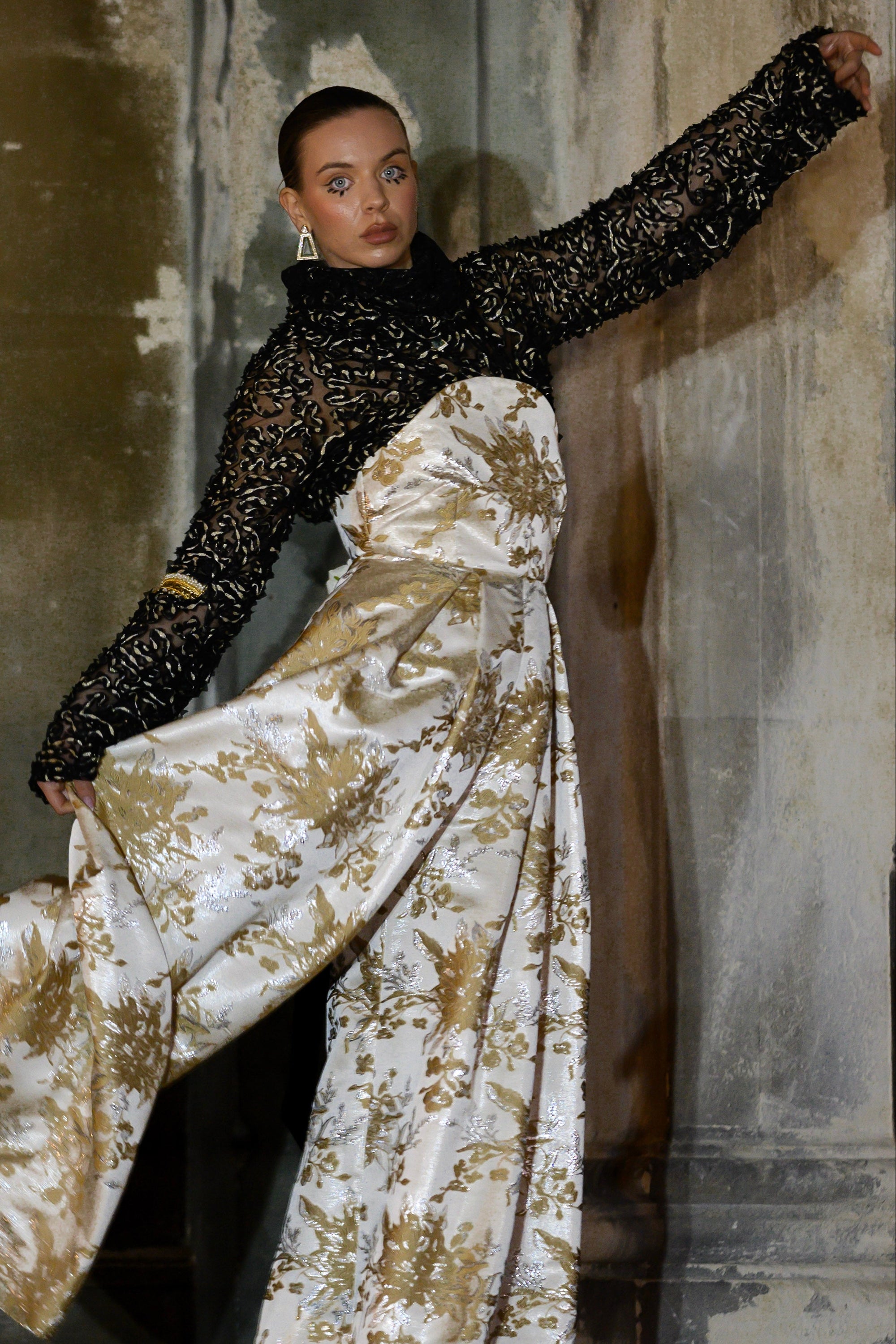 Model wearing a black sequined top and white floral pants against a textured wall.