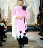 Model wearing a pink and black floral dress on a runway with white draped curtains in the background.