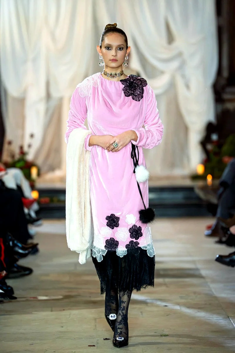 Model wearing a pink and black floral dress on a runway with white draped curtains in the background.