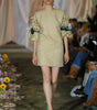 Model walking on a runway wearing a light-colored dress with green accents.