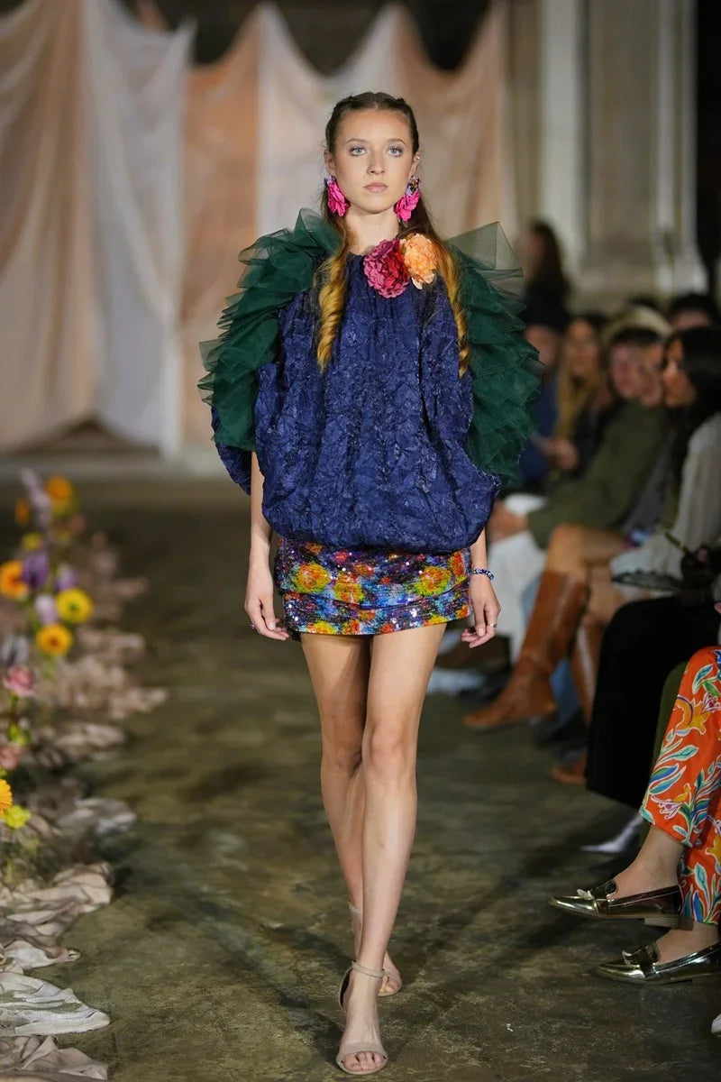 Model walking on a runway wearing a colorful outfit with floral elements.