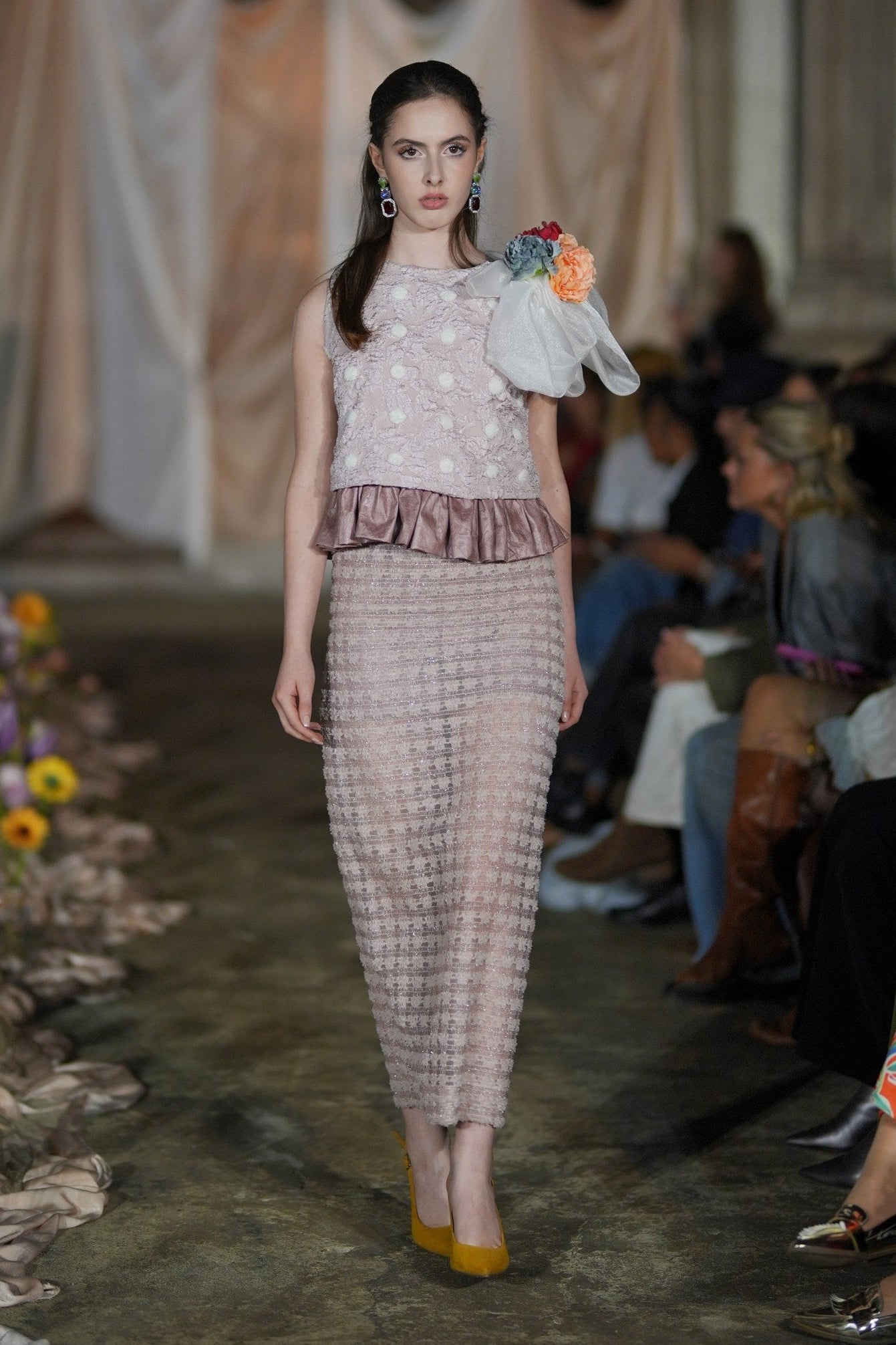 Model walking on a fashion show runway with floral decorations.