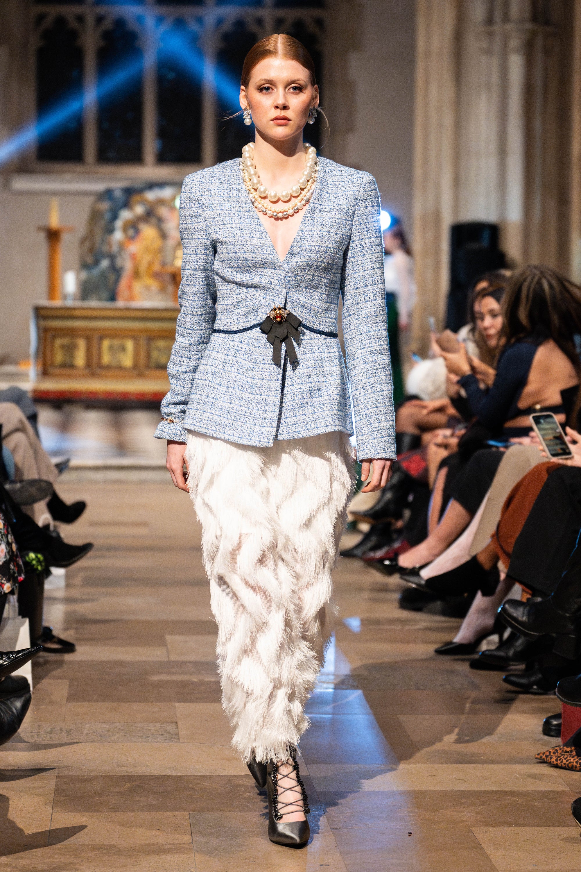 Model walking on a runway wearing a blue checkered blazer and white feathered skirt.