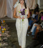 Model walking down a runway in a white dress with a floral headpiece and bouquet.