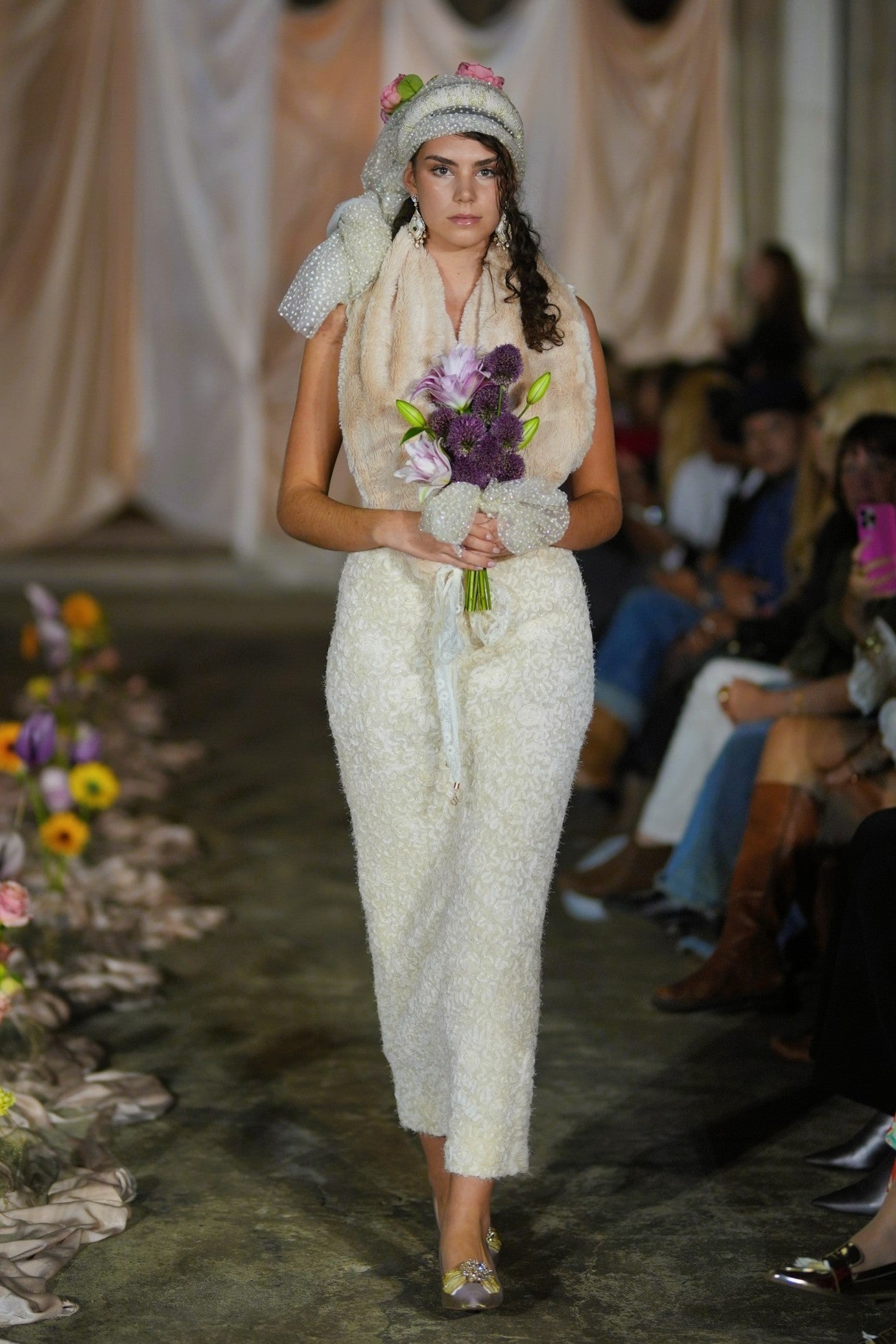 Model walking down a runway in a white dress with a floral headpiece and bouquet.