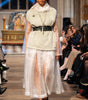 Model walking on a runway wearing a textured top and sheer skirt with a blurred audience in the background.
