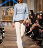Model walking on a runway wearing a blue top and white pants in an indoor setting.
