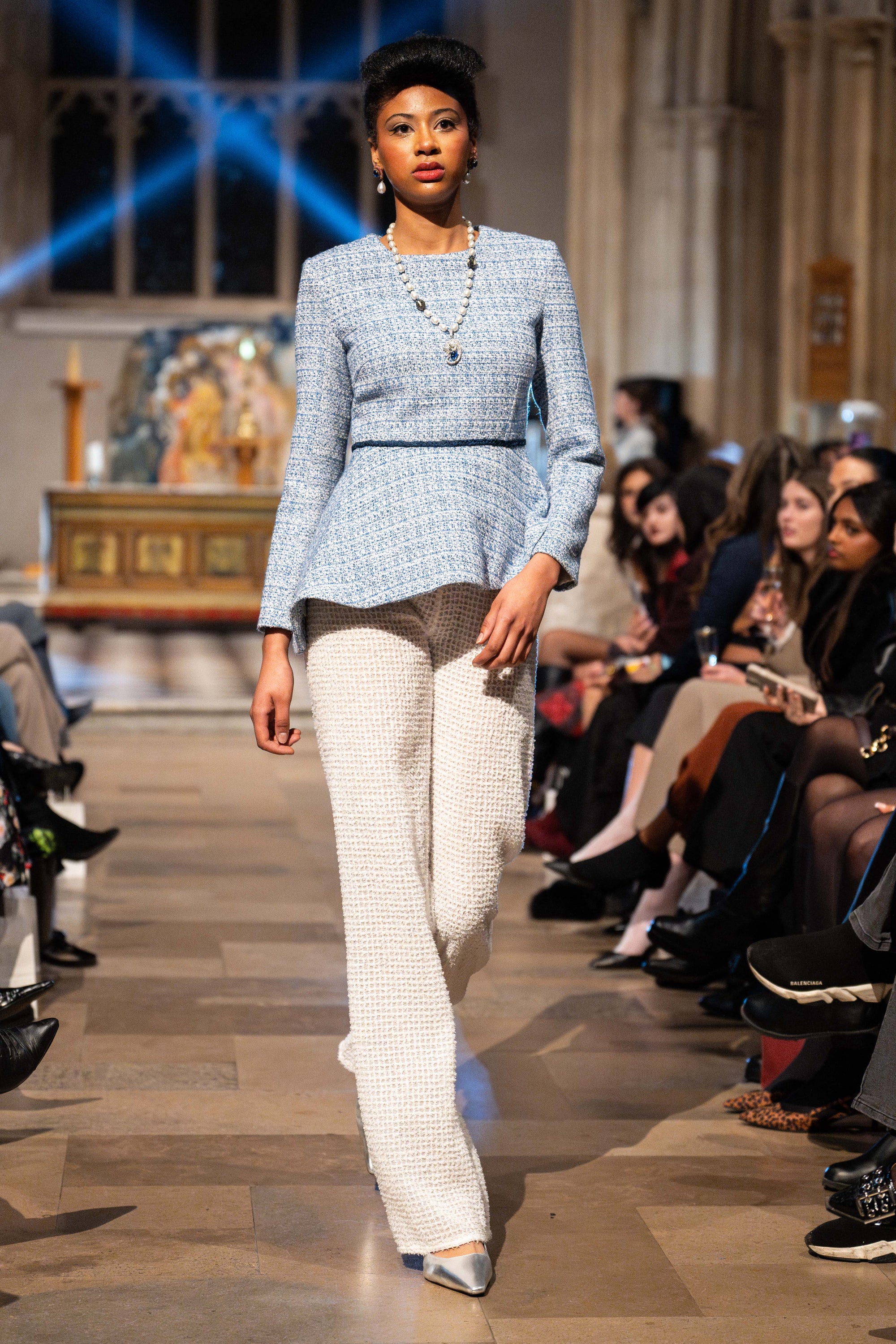 Model walking on a runway wearing a blue top and white pants in an indoor setting.